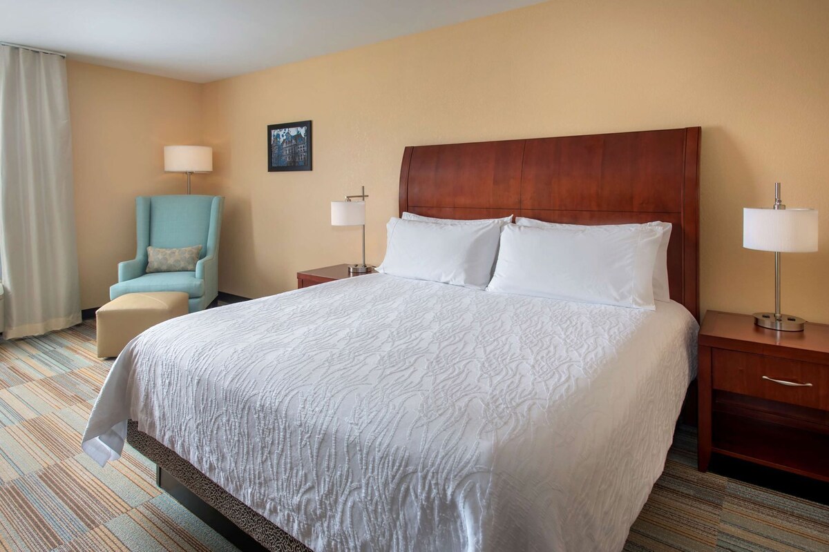 Quarto de hotel com uma cama grande, uma cadeira, duas mesas de cabeceira com luminárias, um quadro na parede e uma janela com cortinas.
