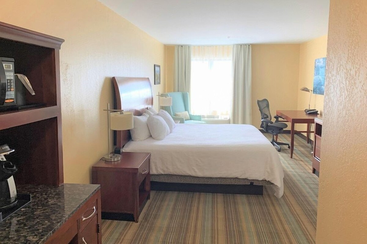 Quarto de hotel com uma cama, escrivaninha, cadeira, abajur, cafeteira e televisão.