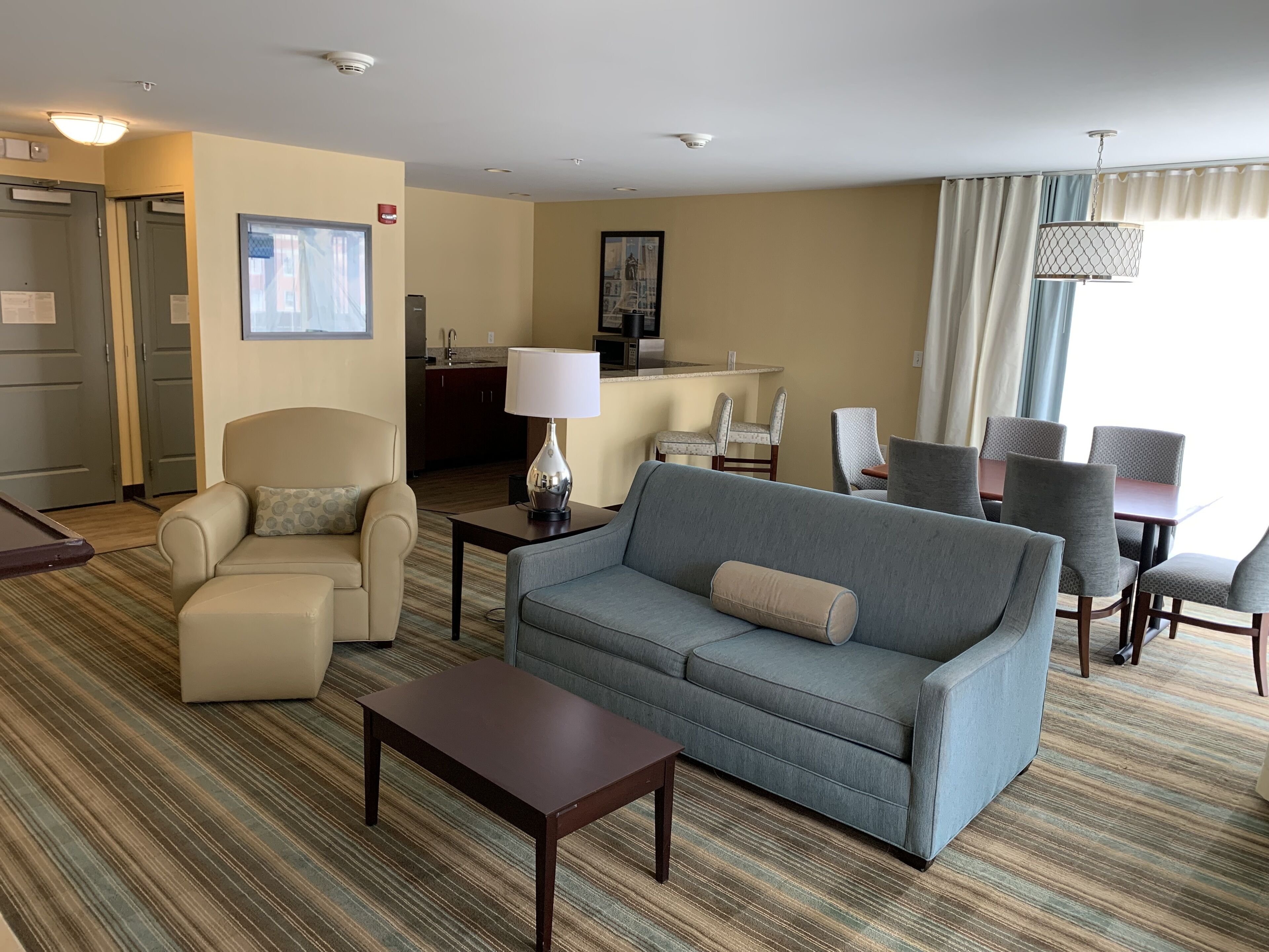 A hotel room with a sofa, armchair, dining table, and chairs. There is a kitchen area in the background.