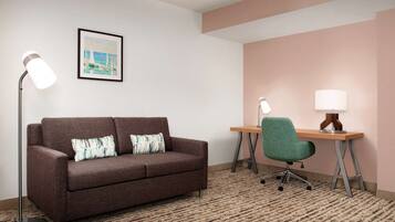 A room with a brown sofa, two lamps, a desk with a chair, and a framed picture on the wall.
