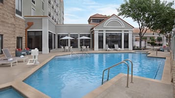 Una piscina con sillas de descanso y sombrillas, una zona de estar techada y un edificio de ladrillo.