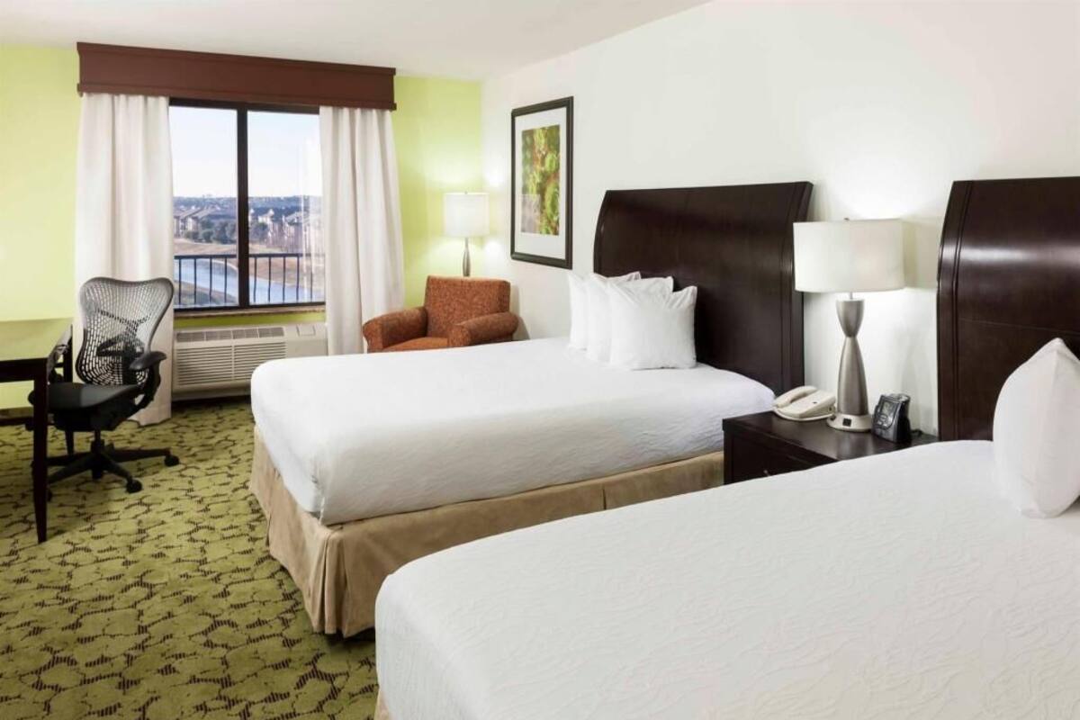 A hotel room with two beds, a desk with a chair, a lamp, a phone, and a window with curtains.