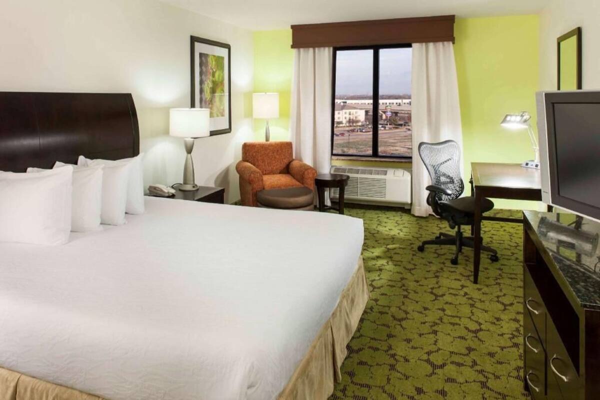 A hotel room with a bed, a desk, a chair, a TV, a window with a view, and a lamp.