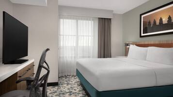 Habitación de hotel con cama, escritorio con televisor, silla, ventana con cortinas y un cuadro en la pared.