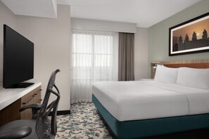 A hotel room with a bed, a desk with a TV, a chair, a window with curtains, and a painting on the wall. - Hilton Garden Inn Dallas Lewisville (Lewisville)