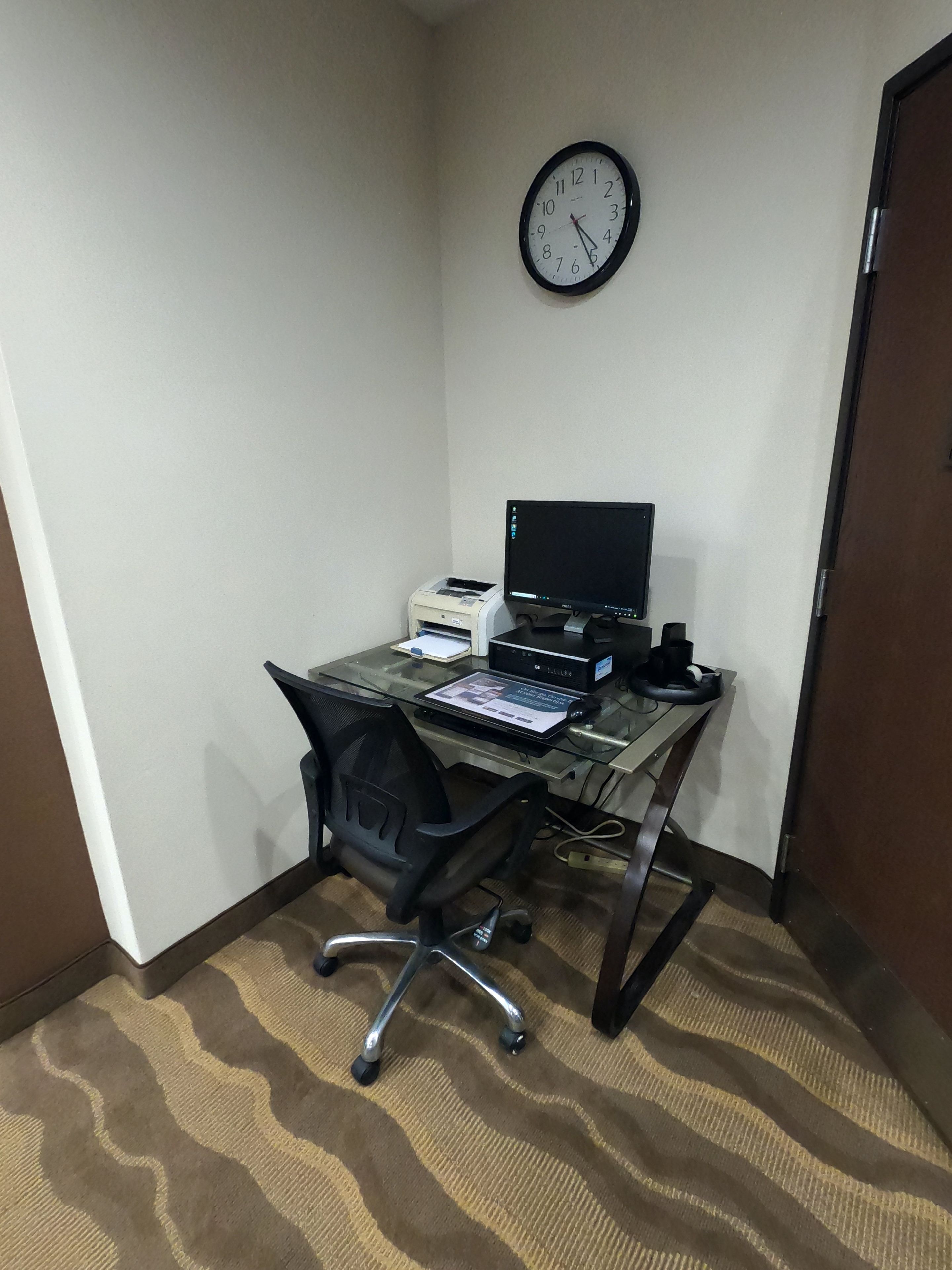 Oficina en esquina con escritorio, computadora, impresora y un reloj en la pared.