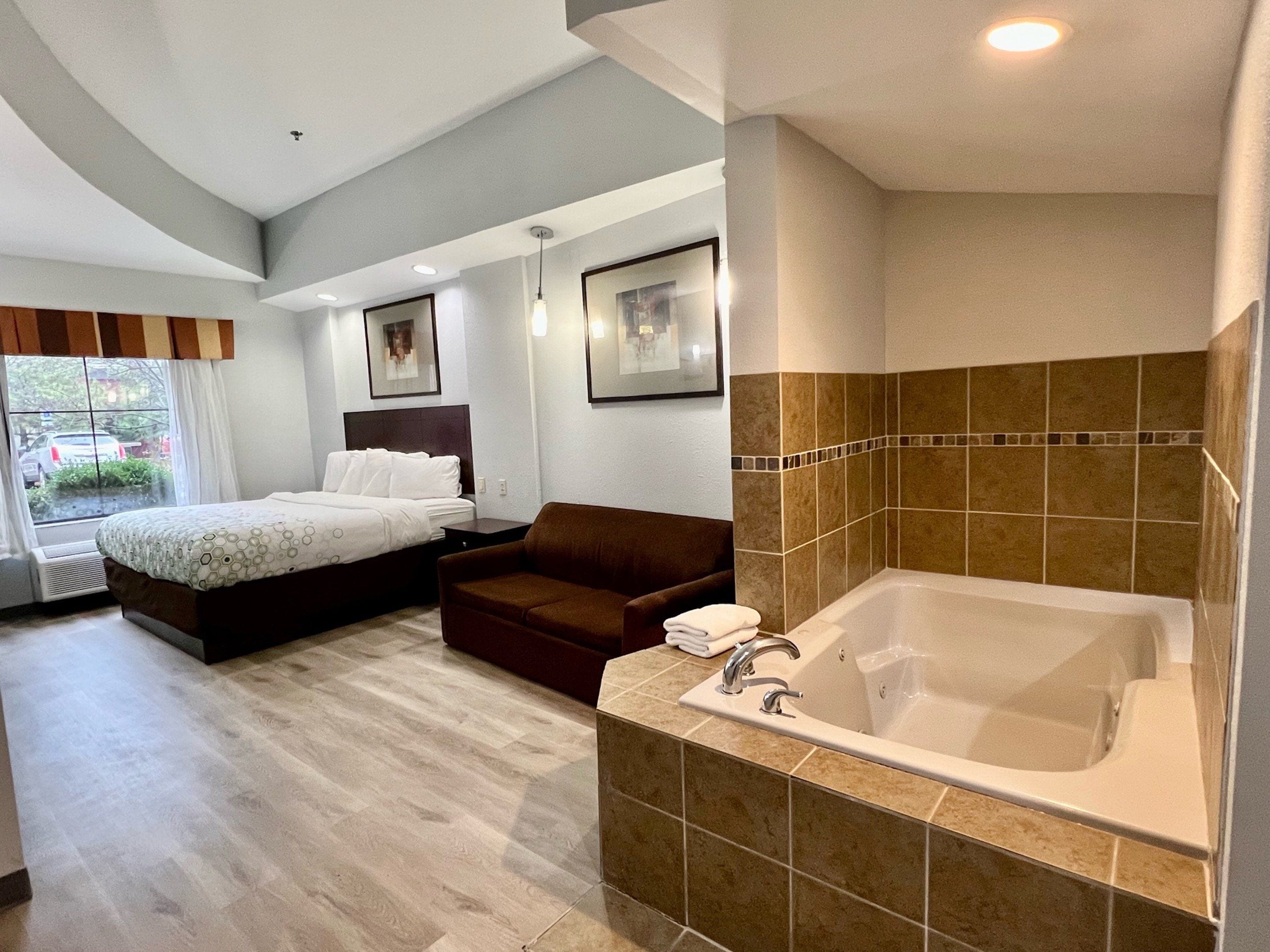 A modern hotel room with a large bed, a sitting area, a bathtub, and a window with a view of the outside.