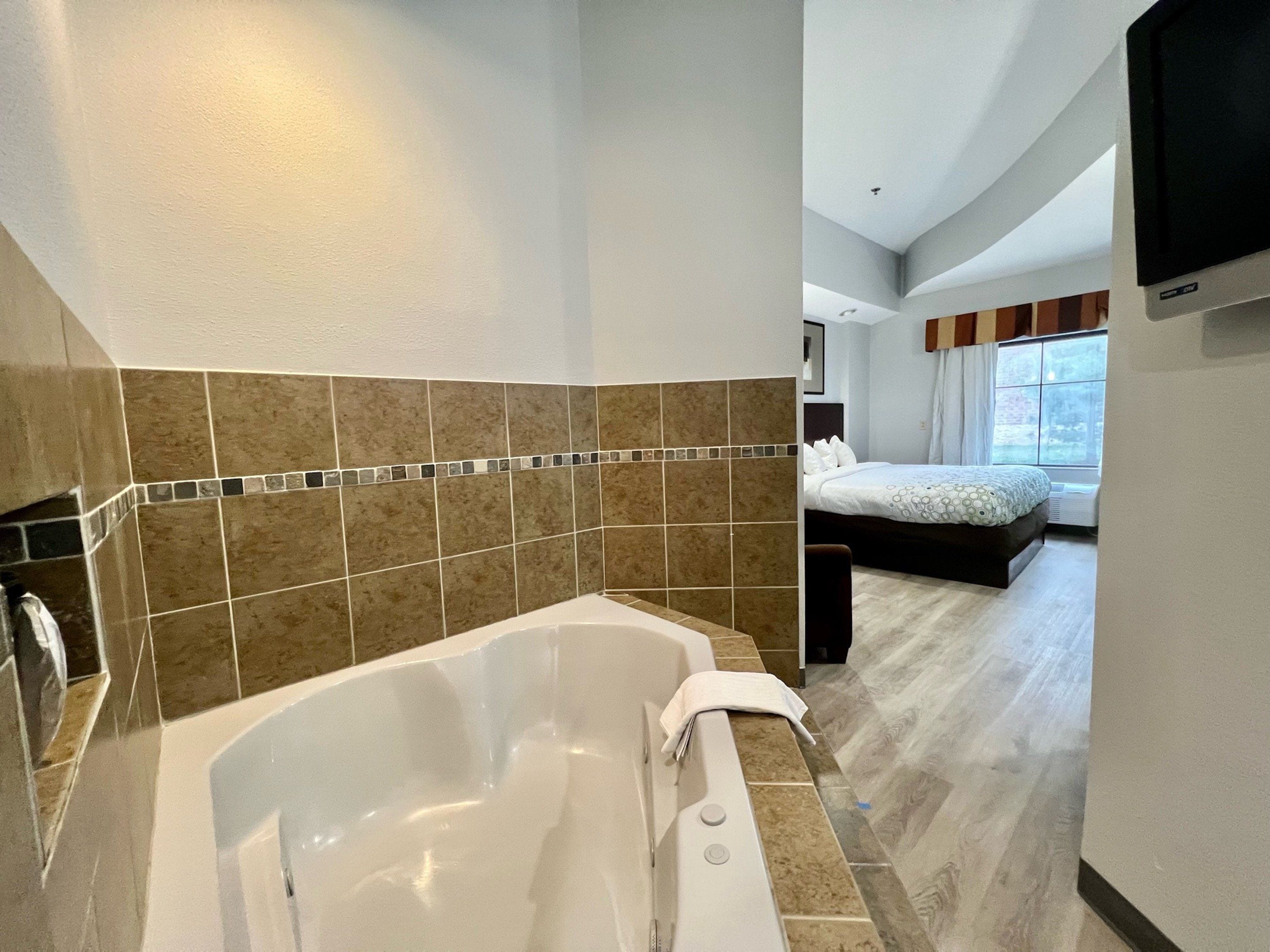 A modern bathroom with a large bathtub, tiled walls, and a separate bedroom with a bed and window.