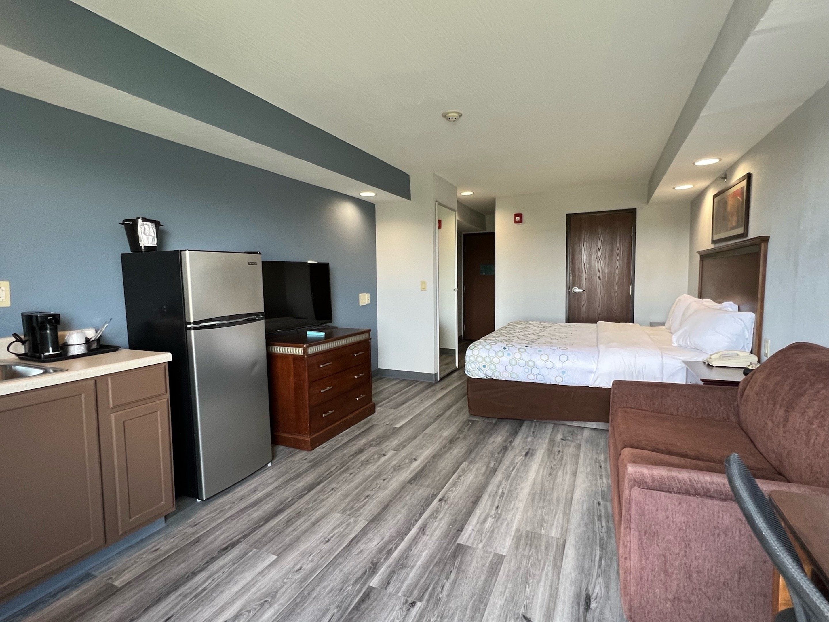 A compact hotel room with a kitchenette, a bed, a sofa, and a wooden floor.