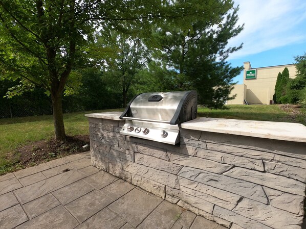 Una parrilla exterior adosada a una pared de piedra, rodeada de césped y árboles.