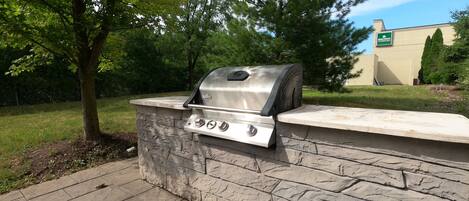 A stone barbecue grill with a metal hood, set against a backdrop of trees and a building with a green sign.