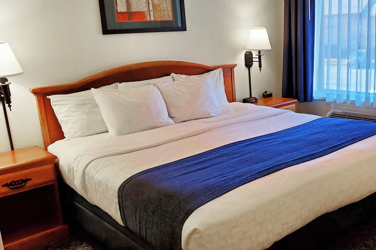 A hotel room with a bed, two bedside tables, a nightstand, a wall-mounted lamp, and a window with curtains.