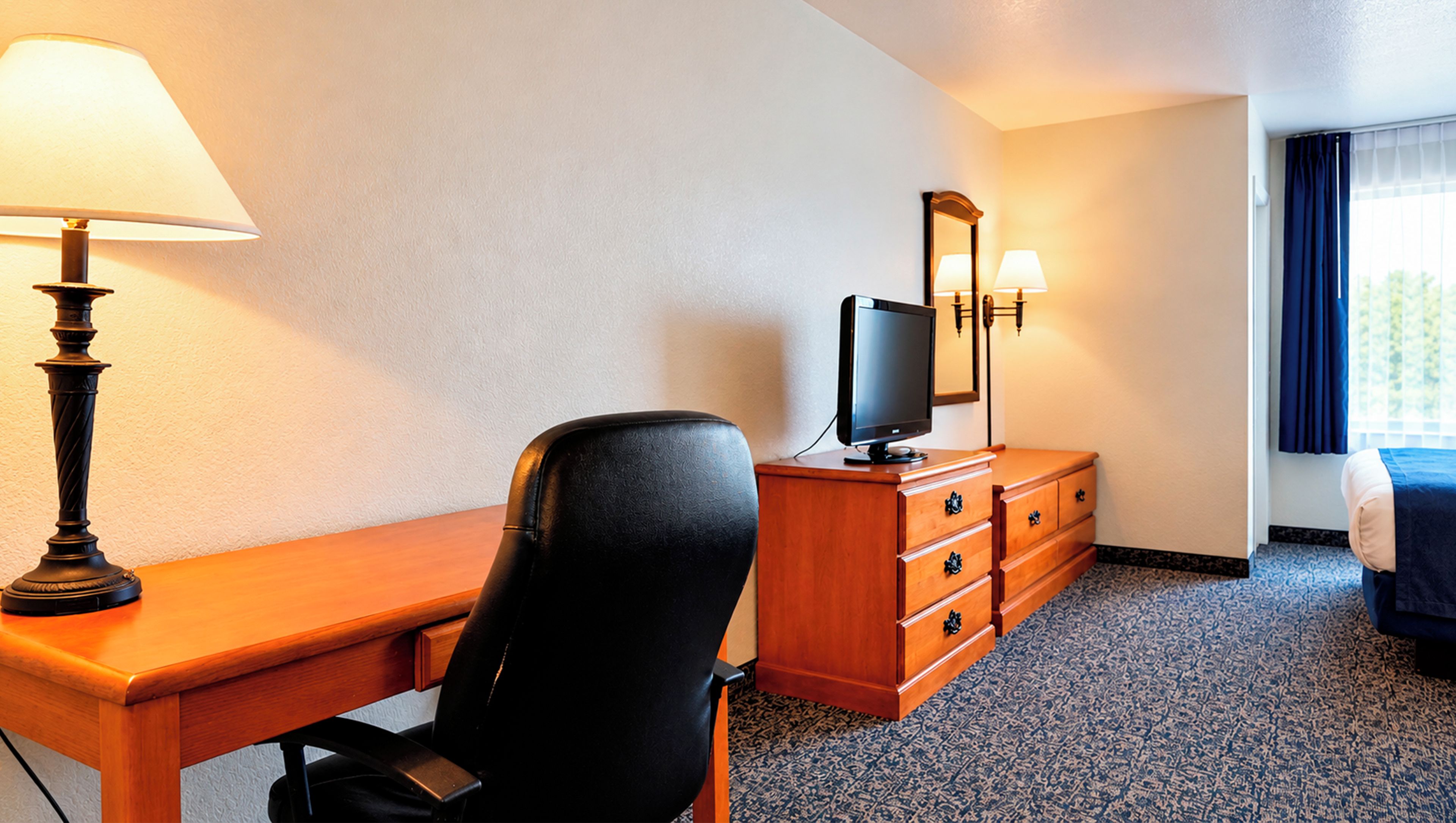 Ein Hotelzimmer mit Schreibtisch, Stuhl, Fernseher, Kommode und einem fensterbepflanzten Fenster.