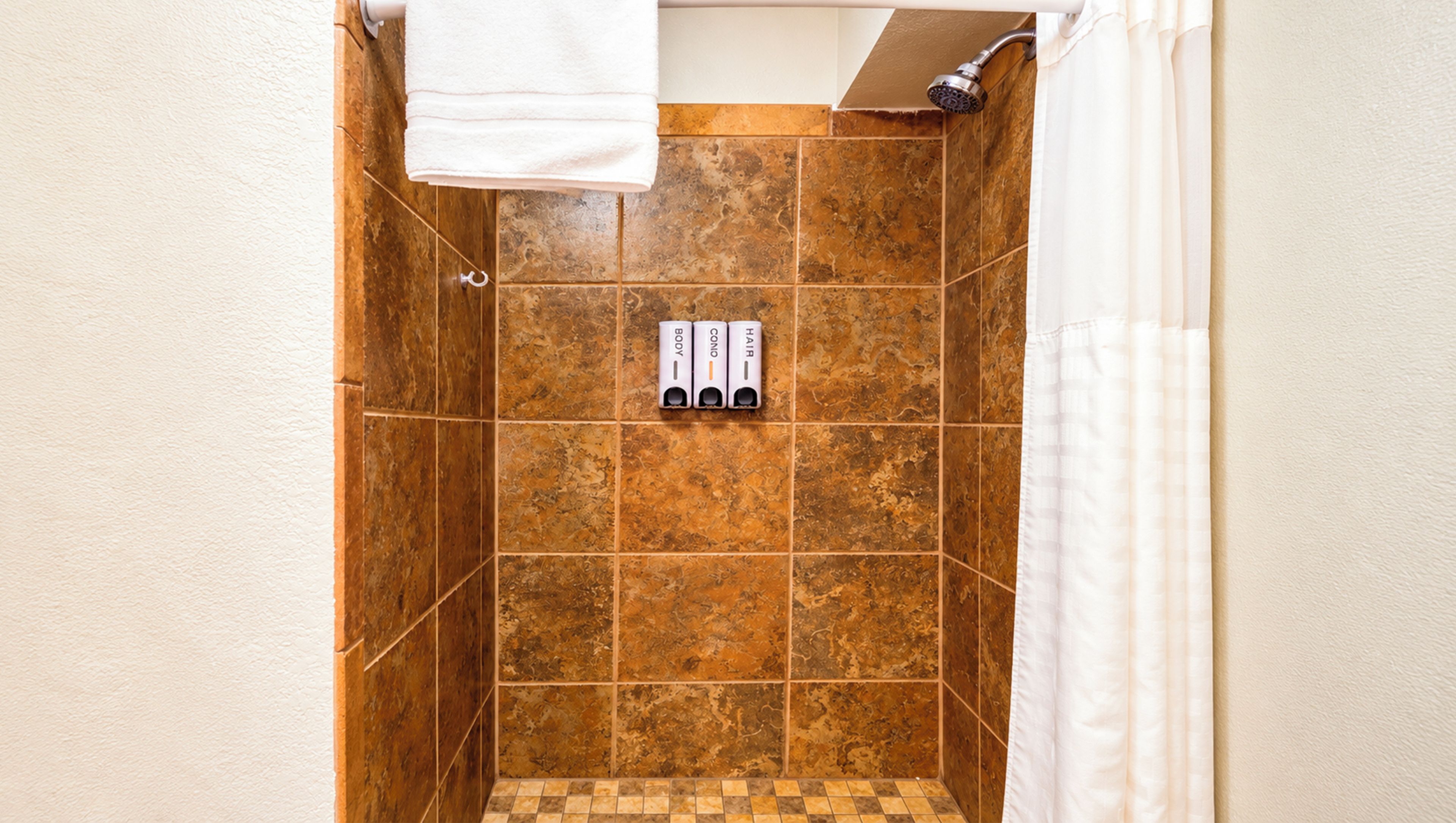 A shower area with brown tiles, a white towel hanging, and three toiletries labeled BODY, CONO, and HAIR.