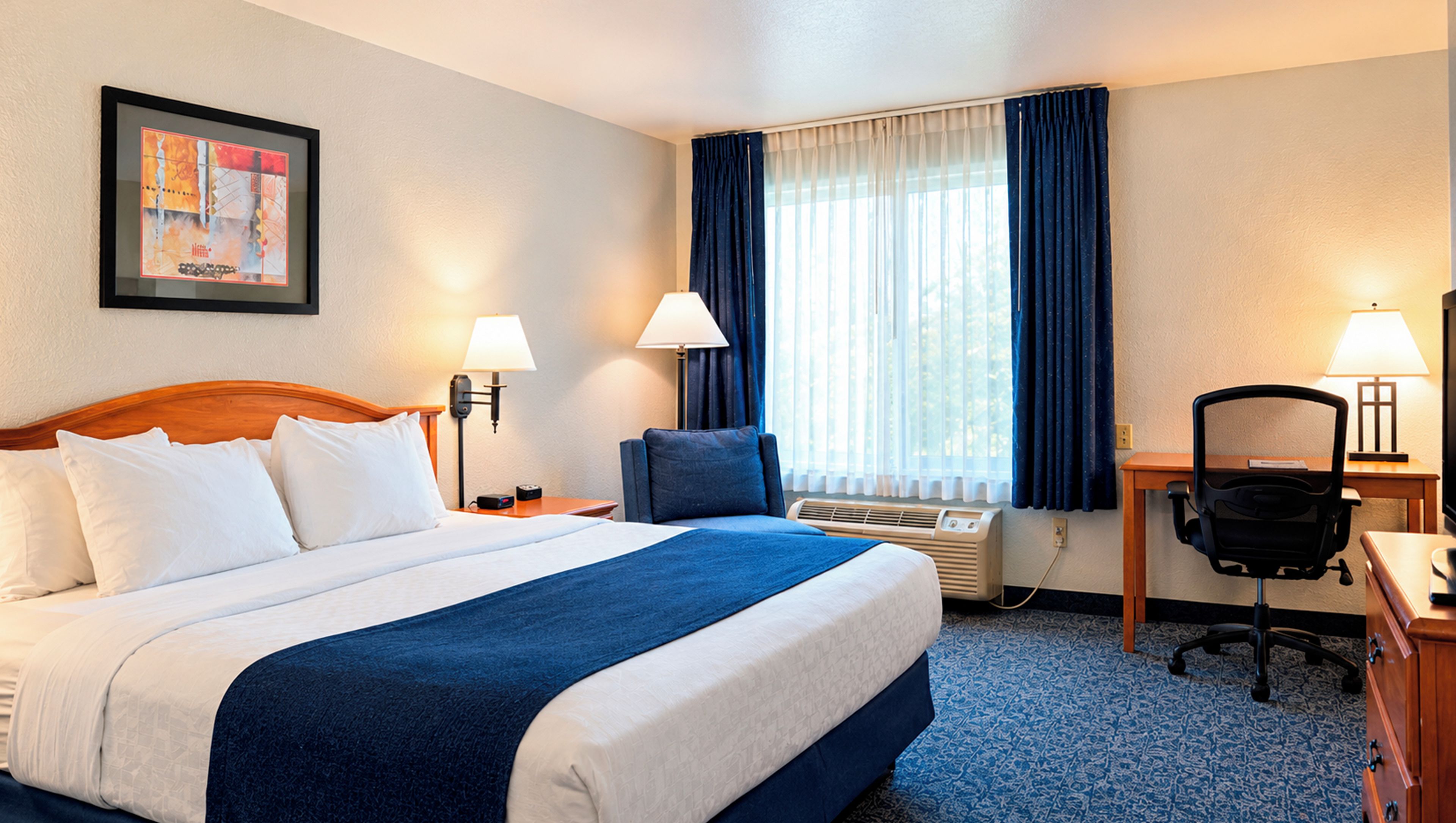 Une chambre d’hôtel comprenant un lit, deux lampes de chevet, un bureau avec une chaise, une fenêtre avec des rideaux et un appareil de climatisation.