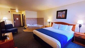 A hotel room with a bed, a desk, a chair, a desk lamp, a wall-mounted TV, a bathroom with a bathtub, and a painting on the wall. - Manitou Pikes Peak Hotel (Manitou Springs)