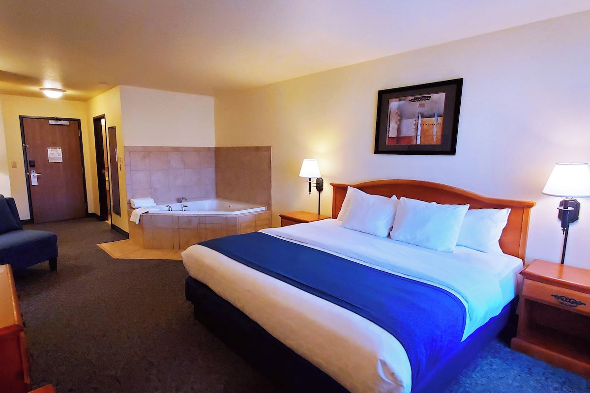 A hotel room with a bed, a desk, a chair, a desk lamp, a wall-mounted TV, a bathroom with a bathtub, and a painting on the wall.