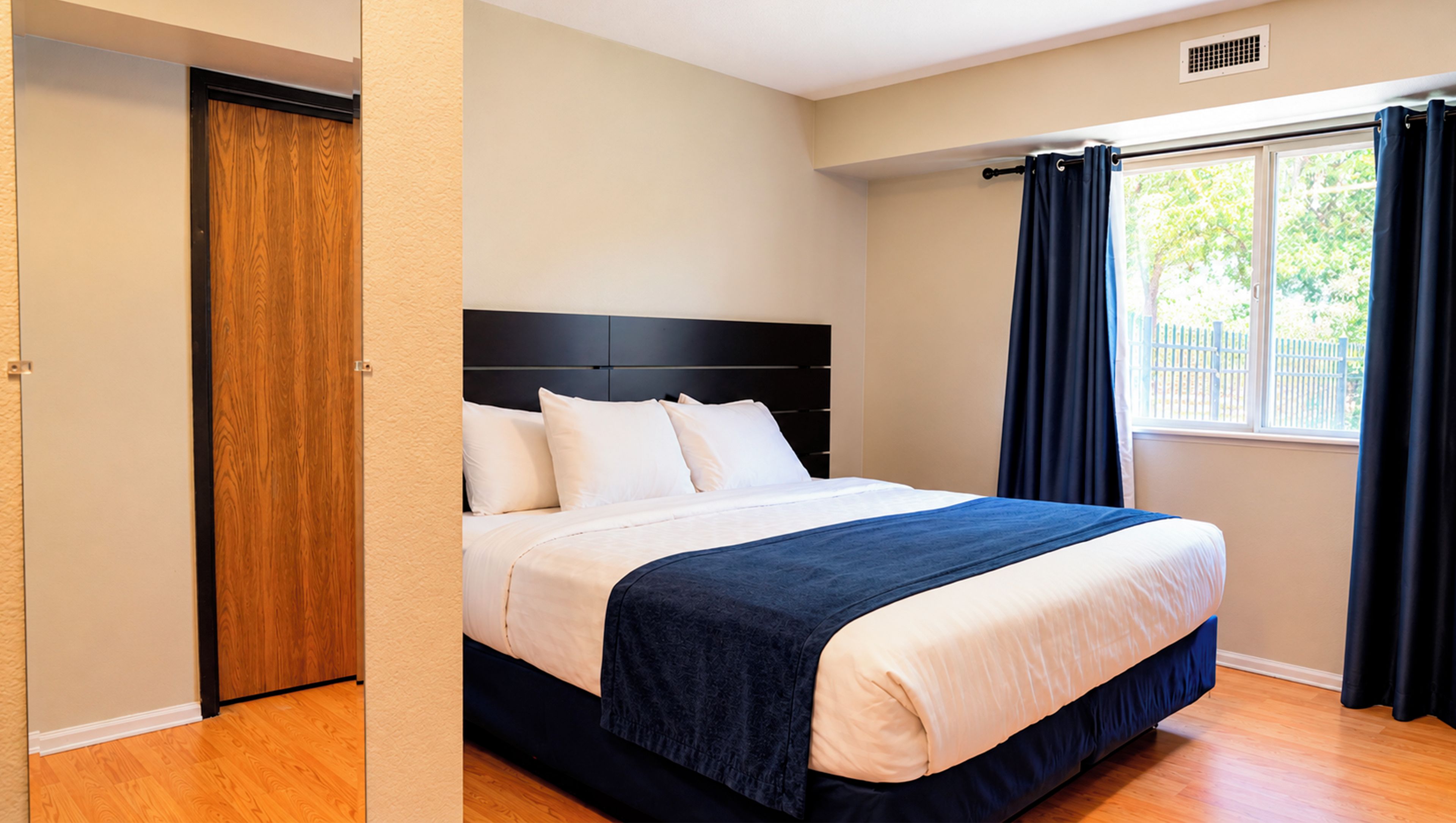 Une chambre avec une porte en bois, un lit avec une literie blanche, une tête de lit sombre et une fenêtre avec des rideaux.