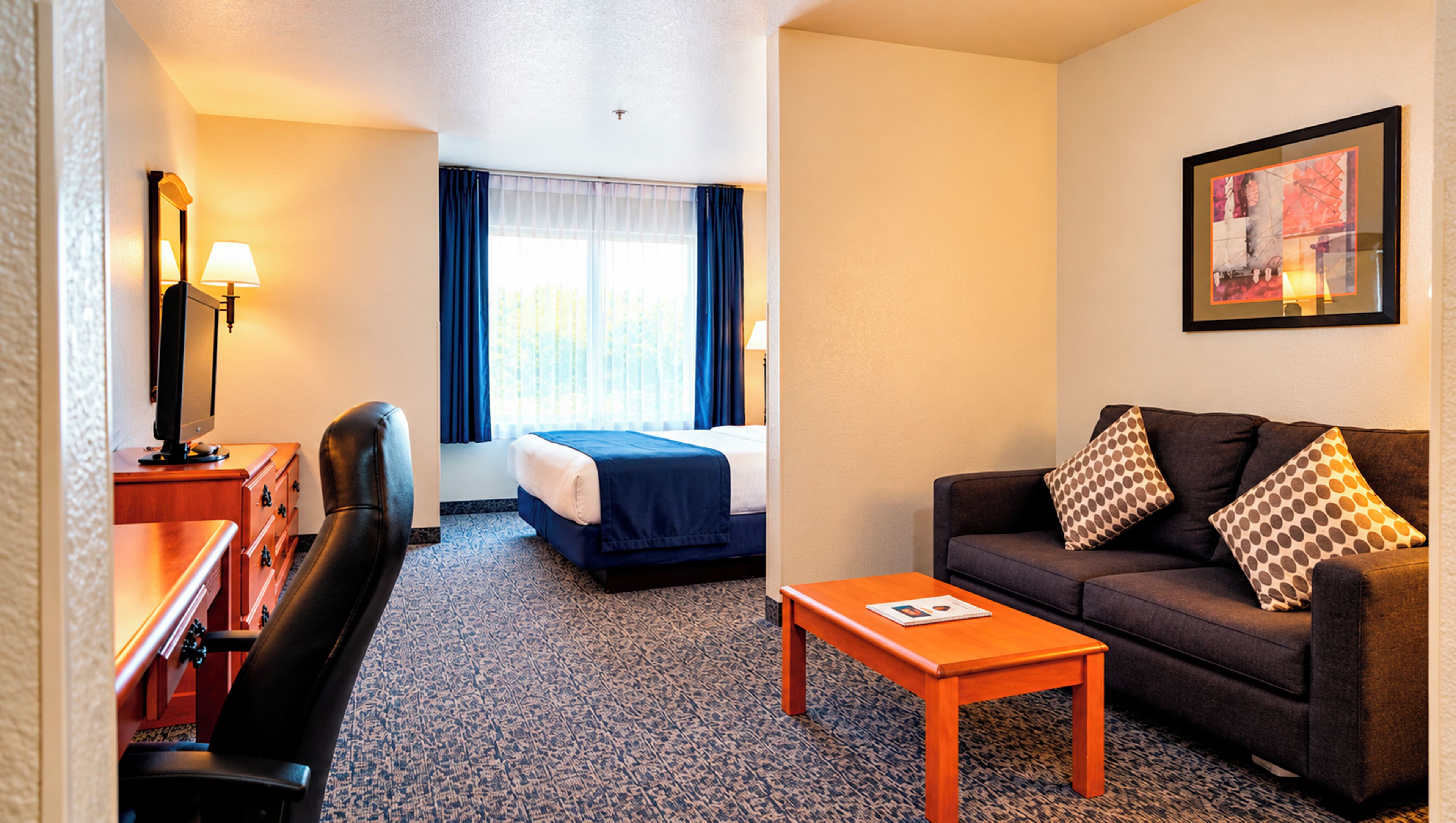 A hotel room with a bed, a sofa, a desk with a lamp, a chair, and a framed picture on the wall.