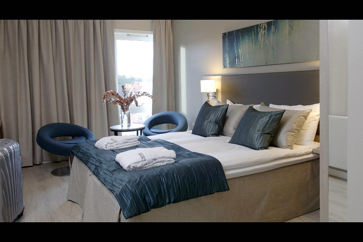 A hotel room with a bed, two chairs, a table, a lamp, and a painting on the wall.