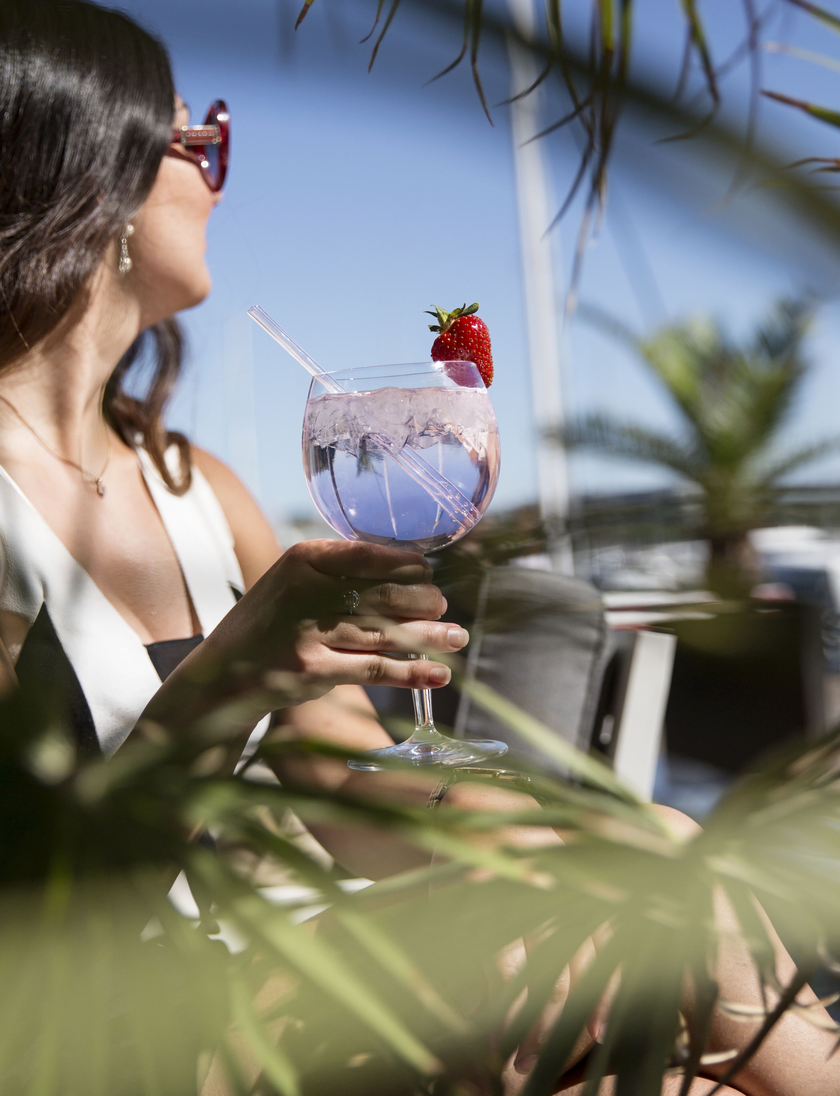 A person holding a cocktail glass with a straw and a strawberry, with a clear view of the sky and palm trees in the background.