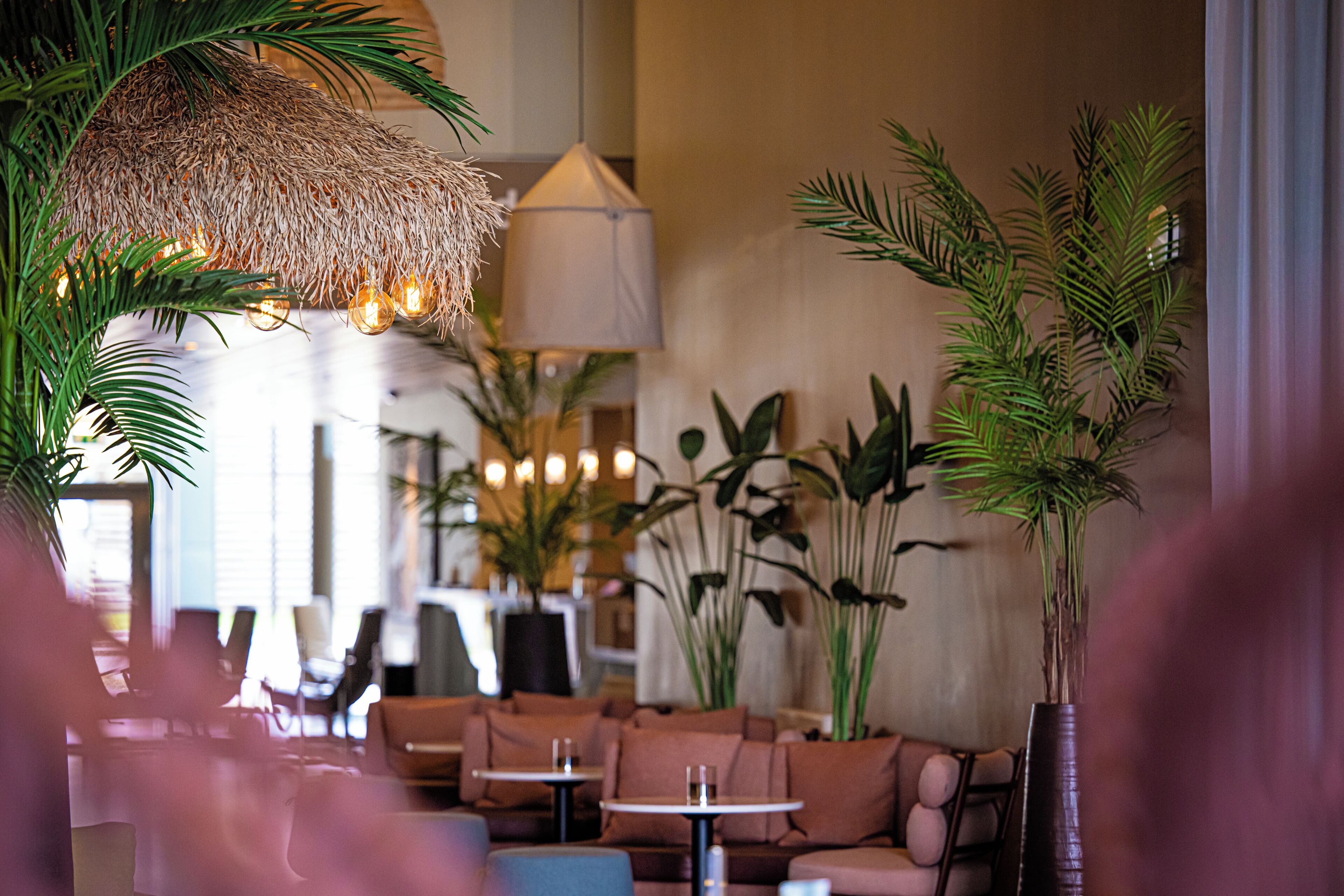 A cozy lounge area with sofas, tables, and hanging lights, featuring tropical plants and a thatched ceiling.