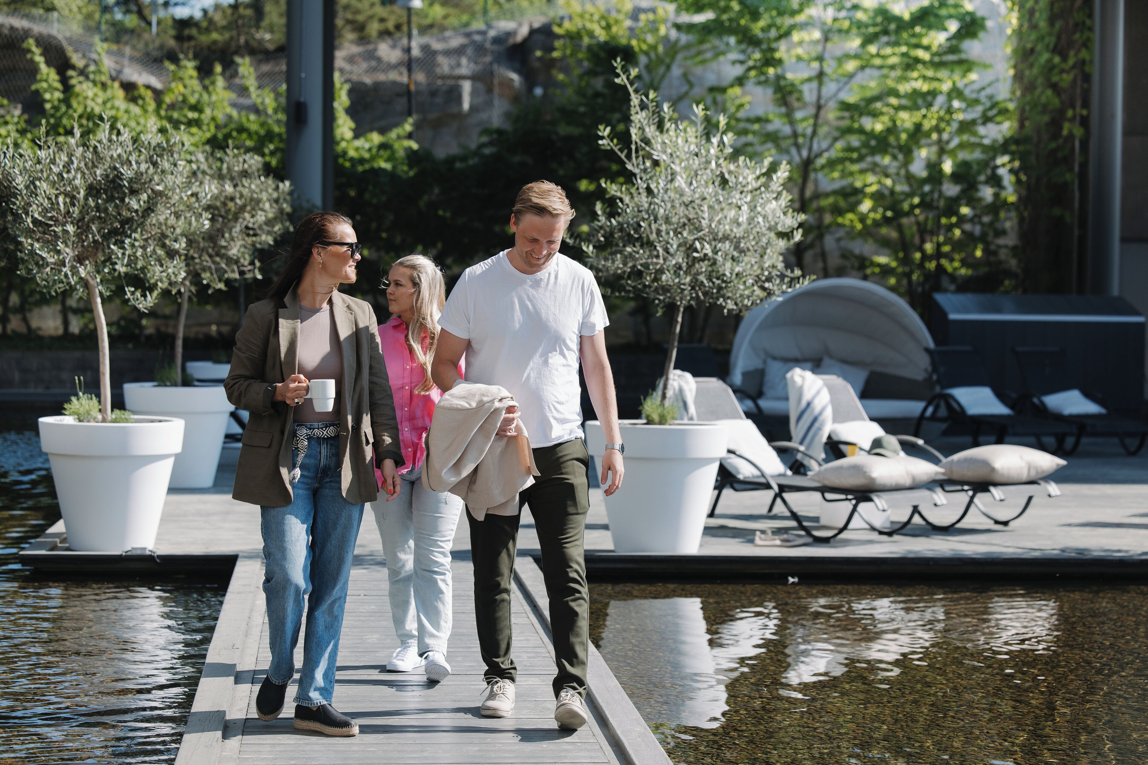 A couple and a woman walking on a wooden deck by a water body, with potted plants and lounge chairs in the background.