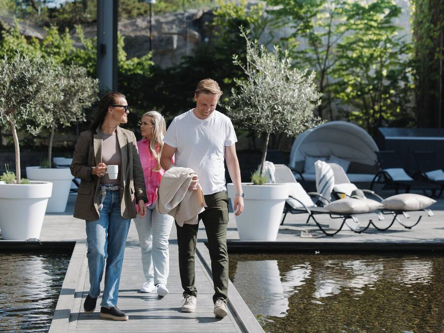 Una pareja y una mujer caminando sobre una cubierta de madera junto a un cuerpo de agua, con plantas en macetas y sillones de descanso al fondo.