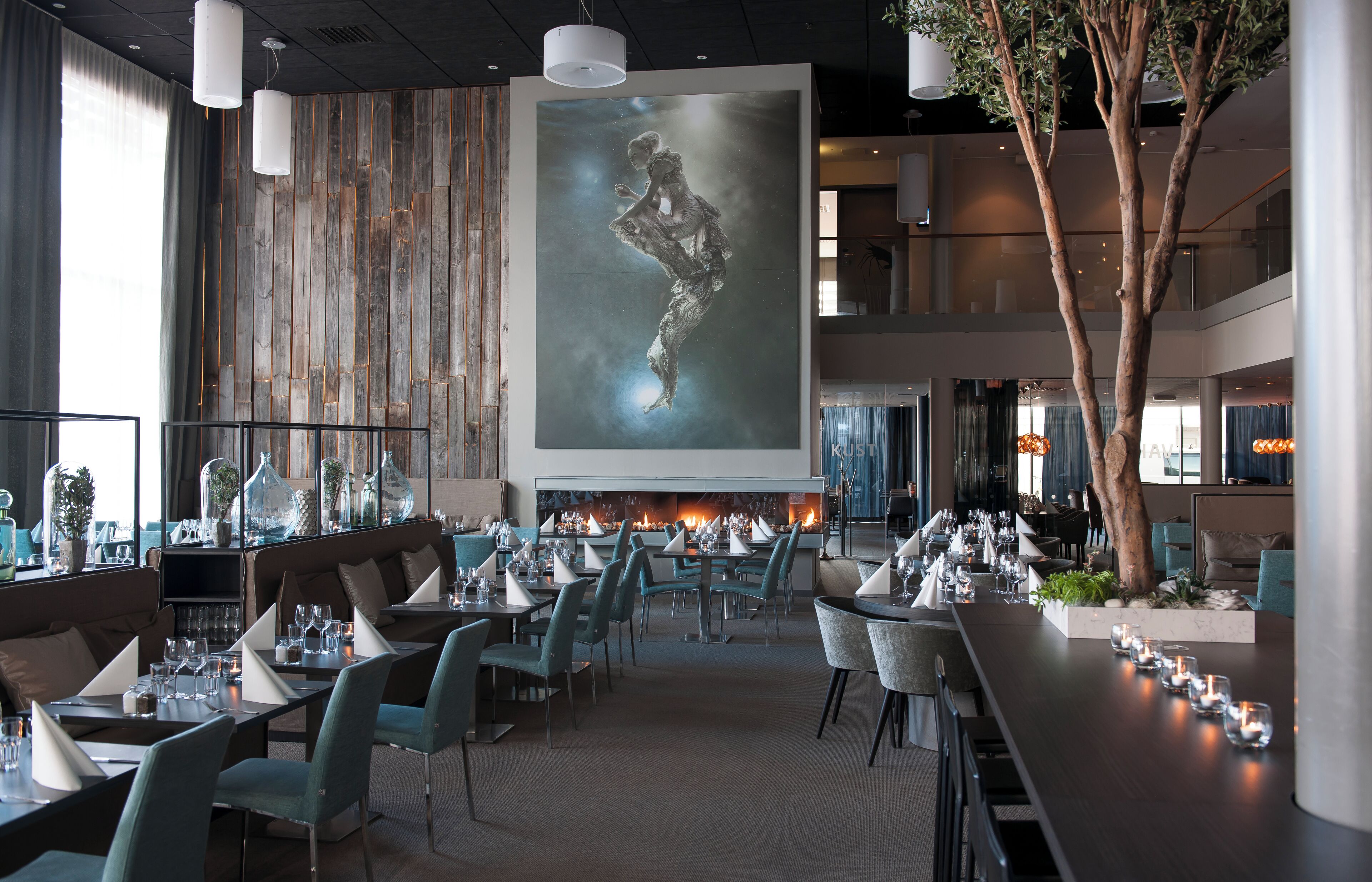 Un restaurant moderne doté d’une cheminée centrale, de plusieurs tables et d’un grand tableau abstrait accroché au mur.