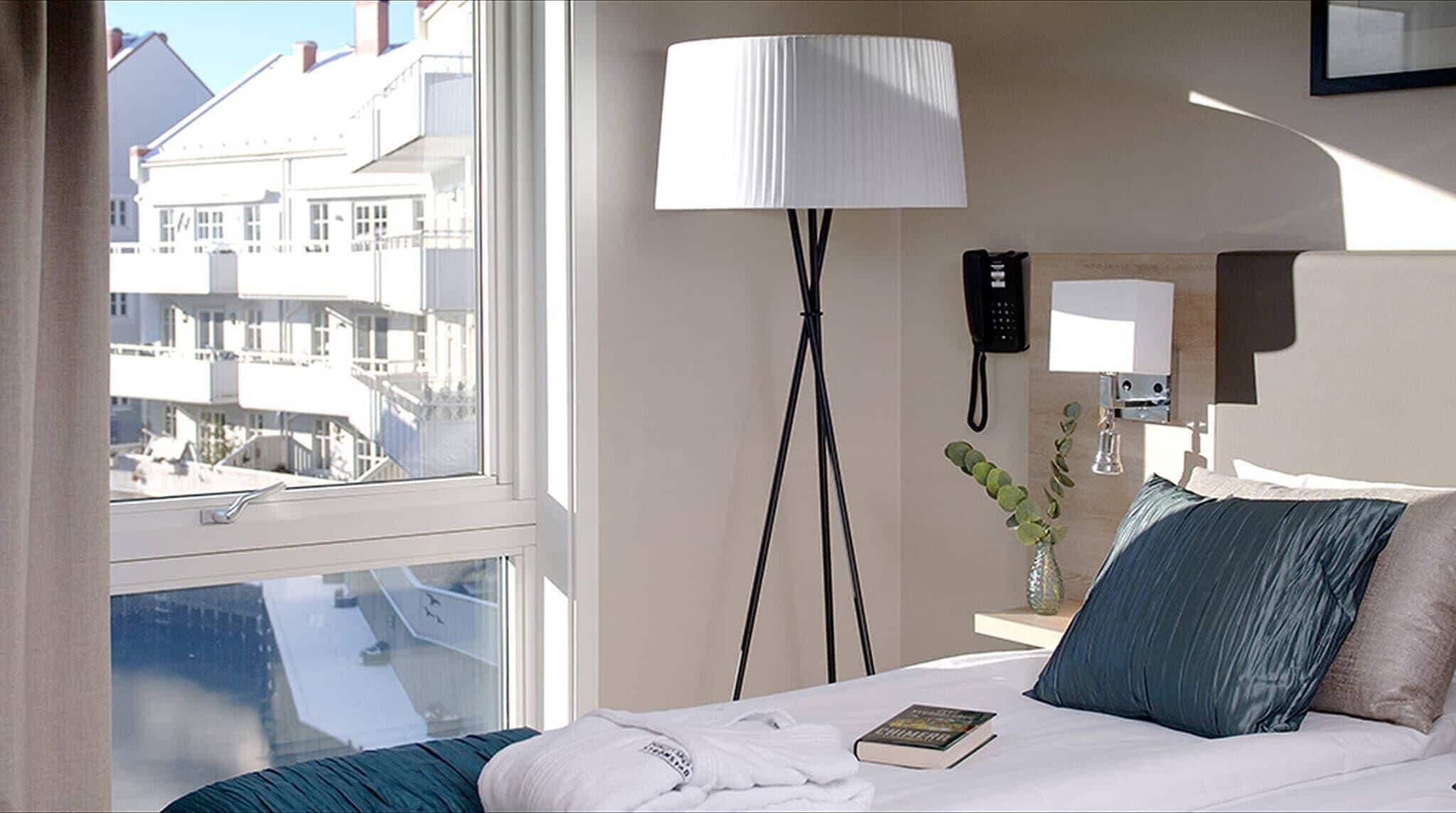 A hotel room with a bed, a bedside lamp, a phone, and a window with a view of buildings.