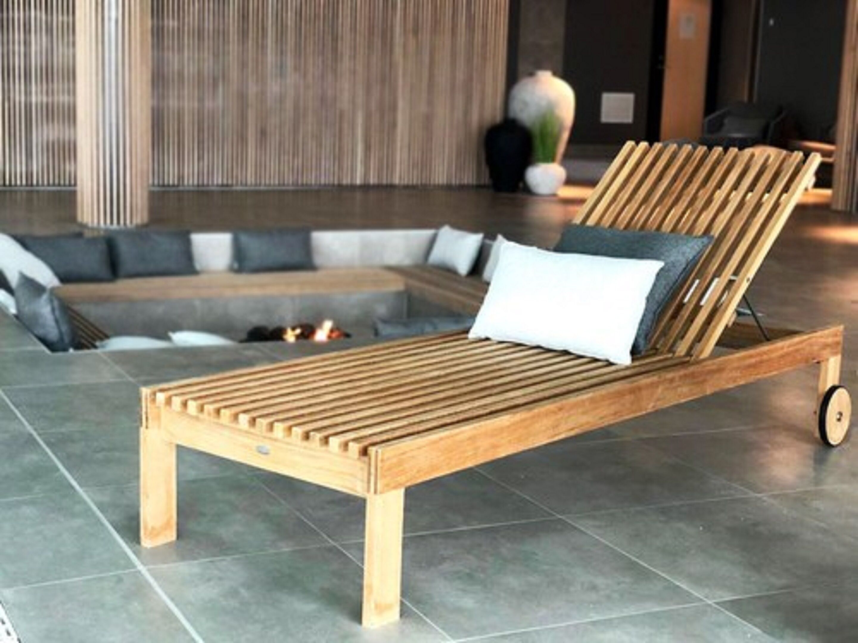 Een houten loungestoel op wielen, een grijze bank en een decoratieve vaas in een moderne lobby.