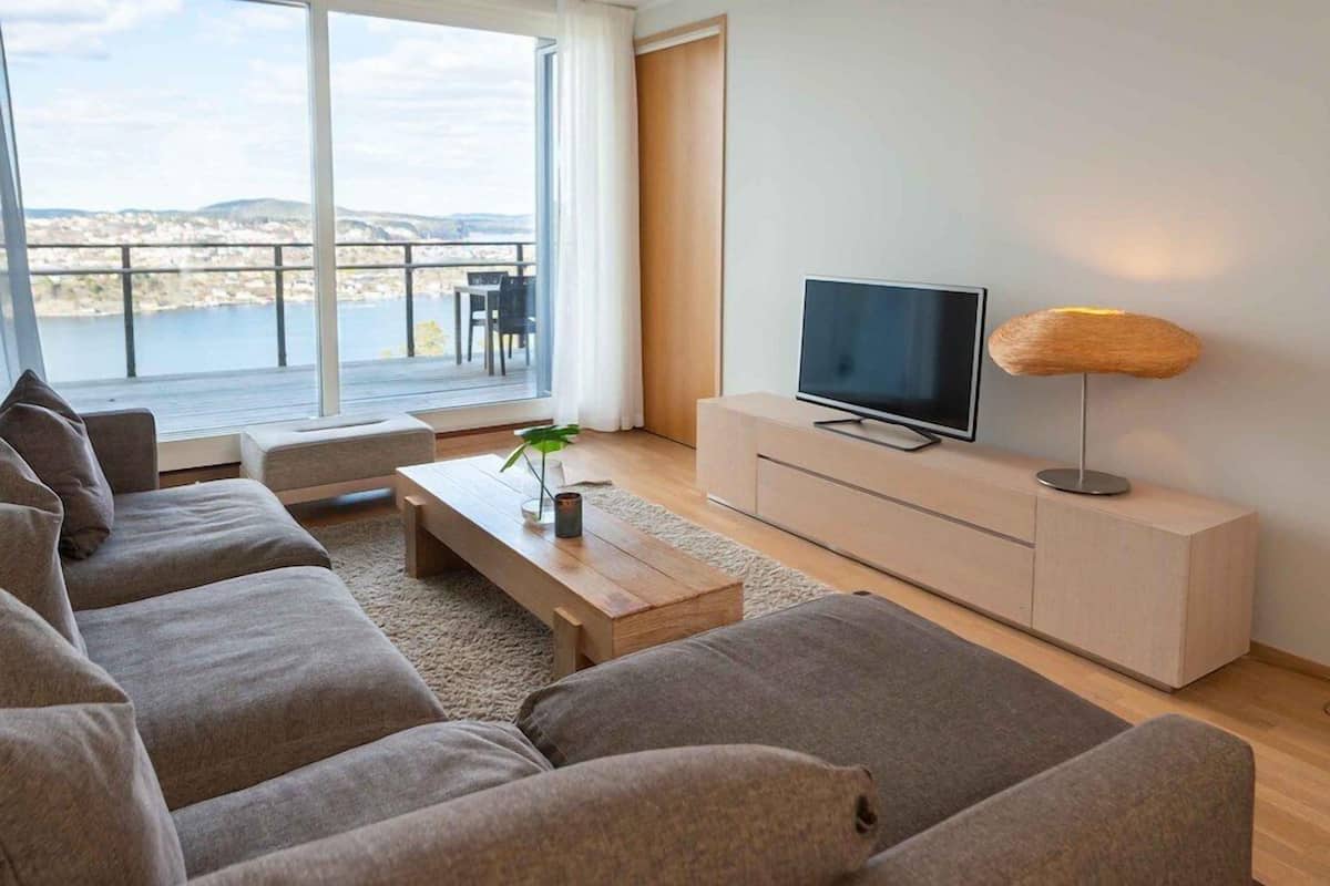 A modern living room with a sofa set, a flat-screen TV, a wooden coffee table, and a view of a body of water through large windows.