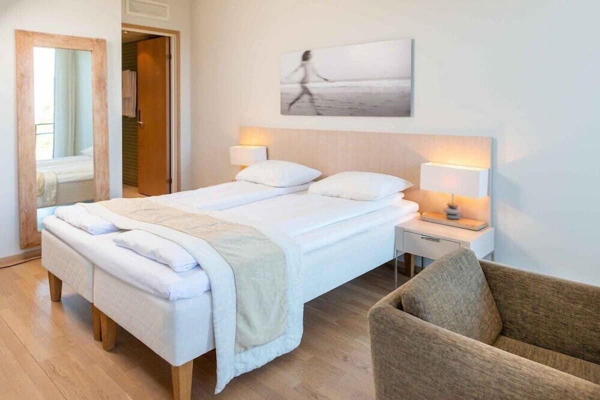 A hotel room with a bed, bedside lamps, a chair, a painting, and a door leading to another room.
