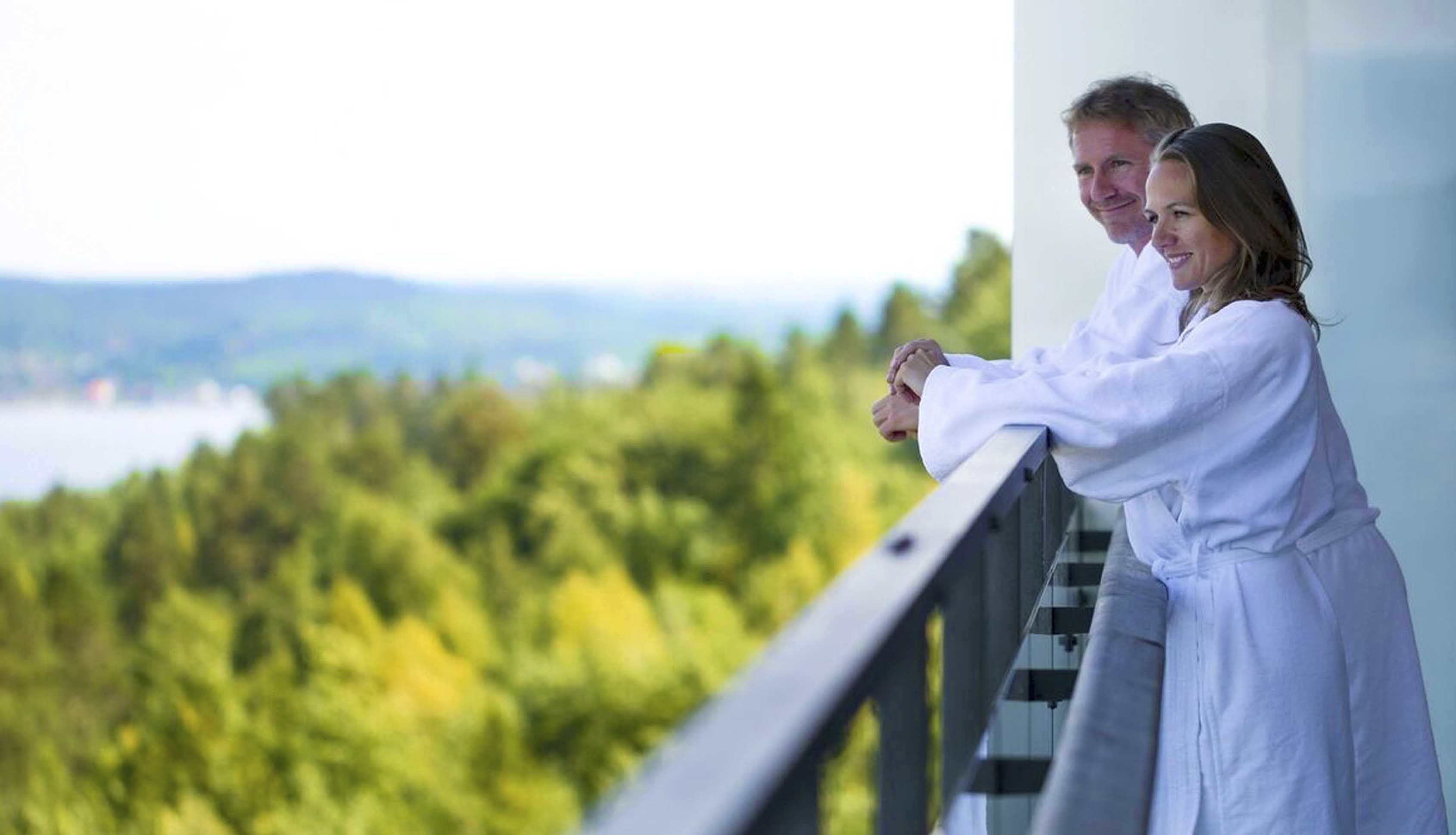 Un couple, vêtu de longues robes blanches, se tient sur un balcon d’où l’on aperçoit un paysage boisé.