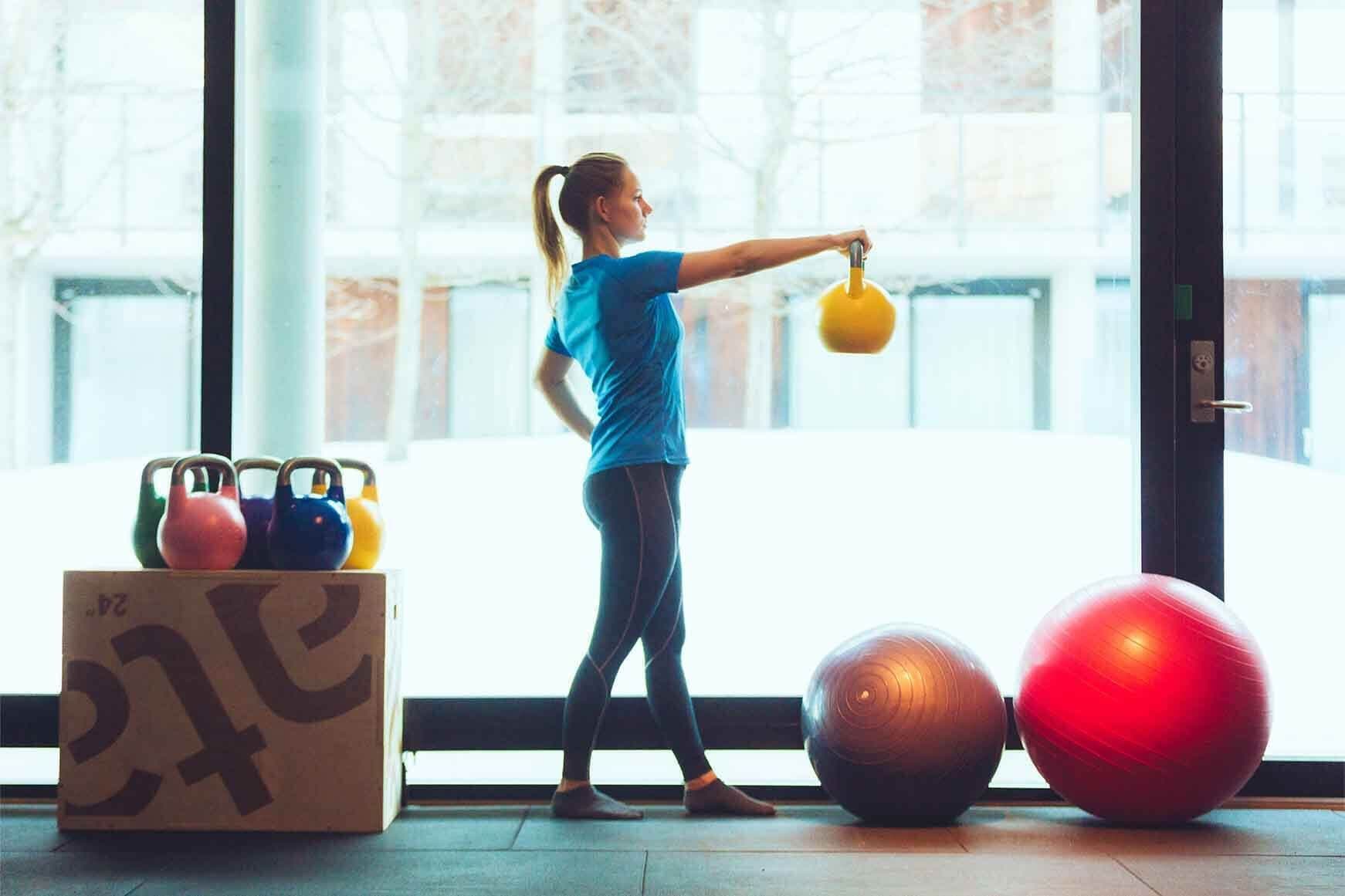 A person exercising with kettlebells and exercise balls in a room with large windows.