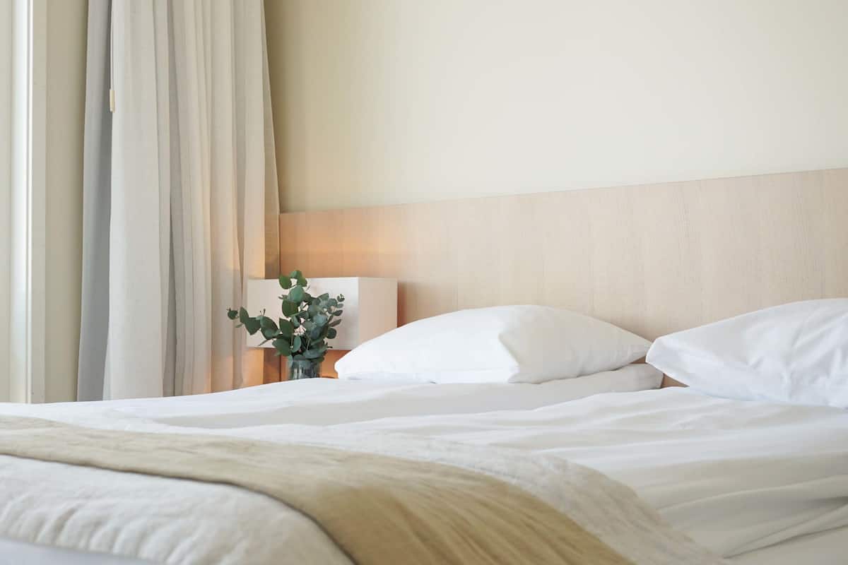 A neatly made bed with white linens, a wooden headboard, and a bedside table with a lamp and a vase of green plants.