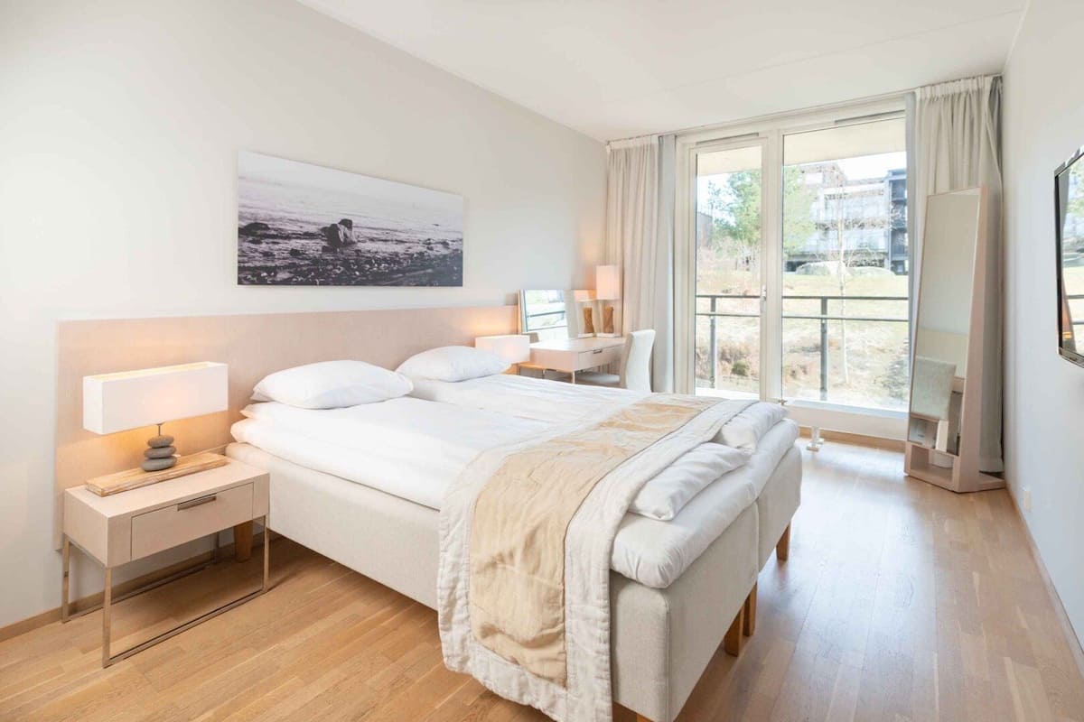 A modern bedroom with a large bed, bedside tables, a nightstand, a window with curtains, and a view of the outdoors.