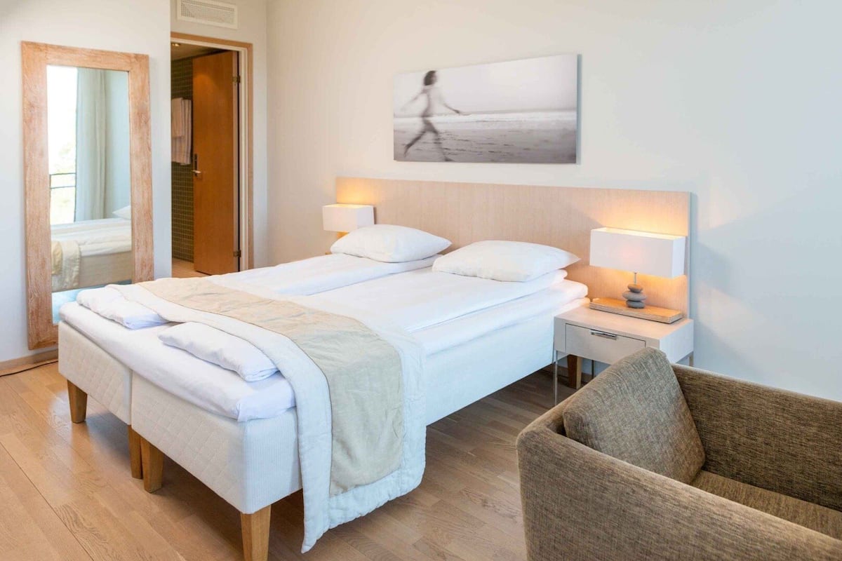 A hotel room with a double bed, bedside tables with lamps, a chair, a large mirror, and a beach-themed artwork on the wall.