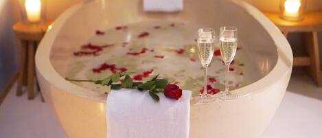 A bathtub with rose petals, two champagne flutes, and a single red rose.