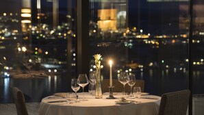 A table set for dining with a candle, glasses, and a vase with a flower, overlooking a cityscape at night.