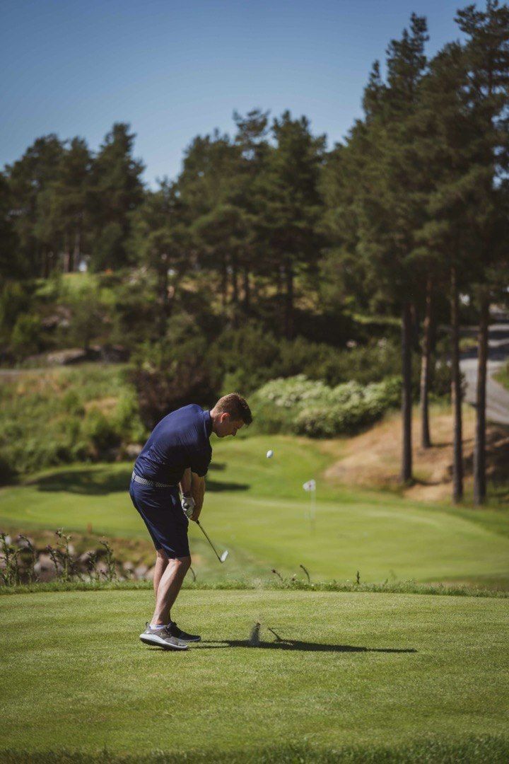 A person playing golf on a well-maintained course with trees and a clear sky in the background.