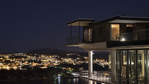 A modern building with a balcony overlooking a lit-up town and a body of water.