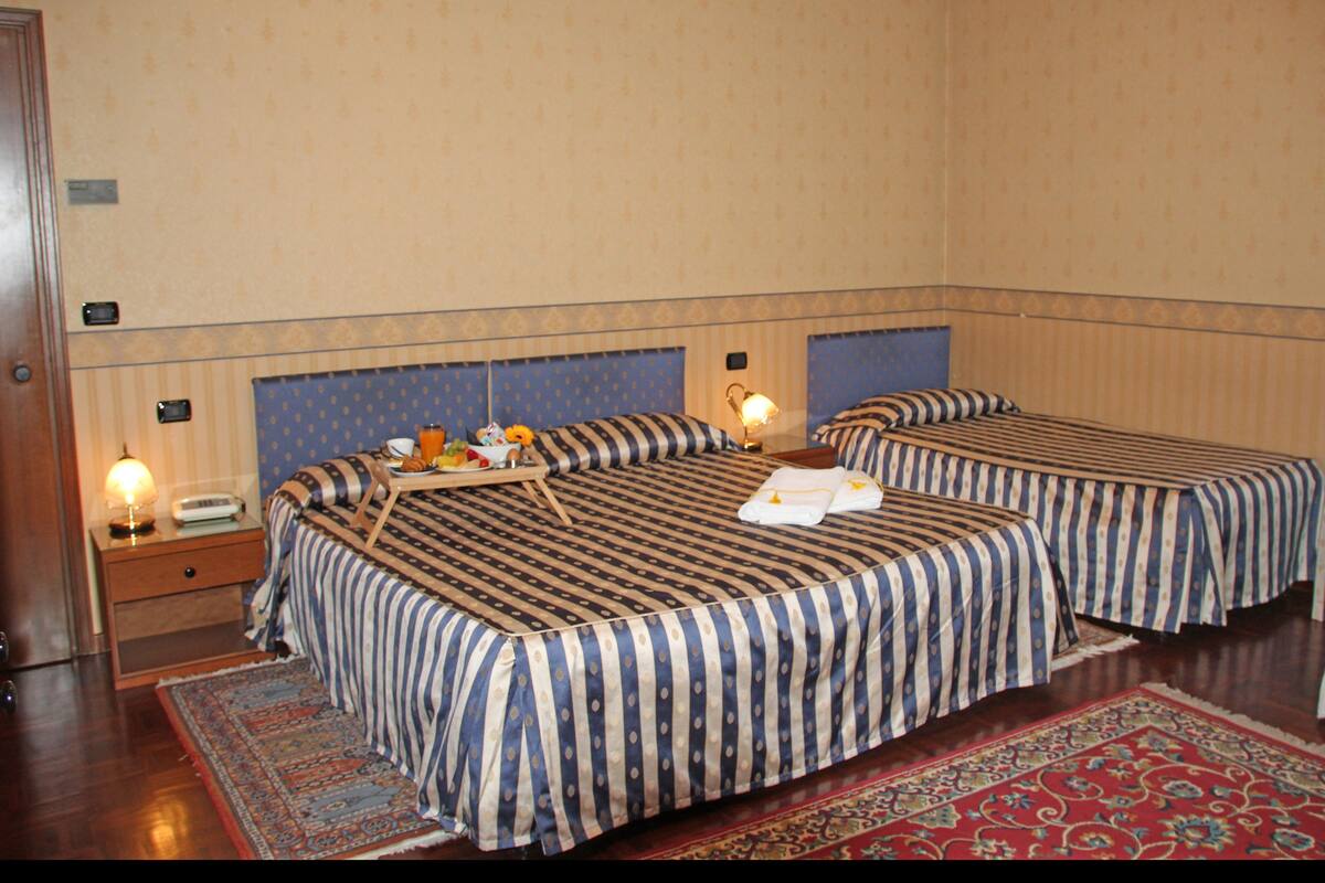 A hotel room with two beds, a wooden nightstand, a bedside table with a lamp, and a red patterned rug.