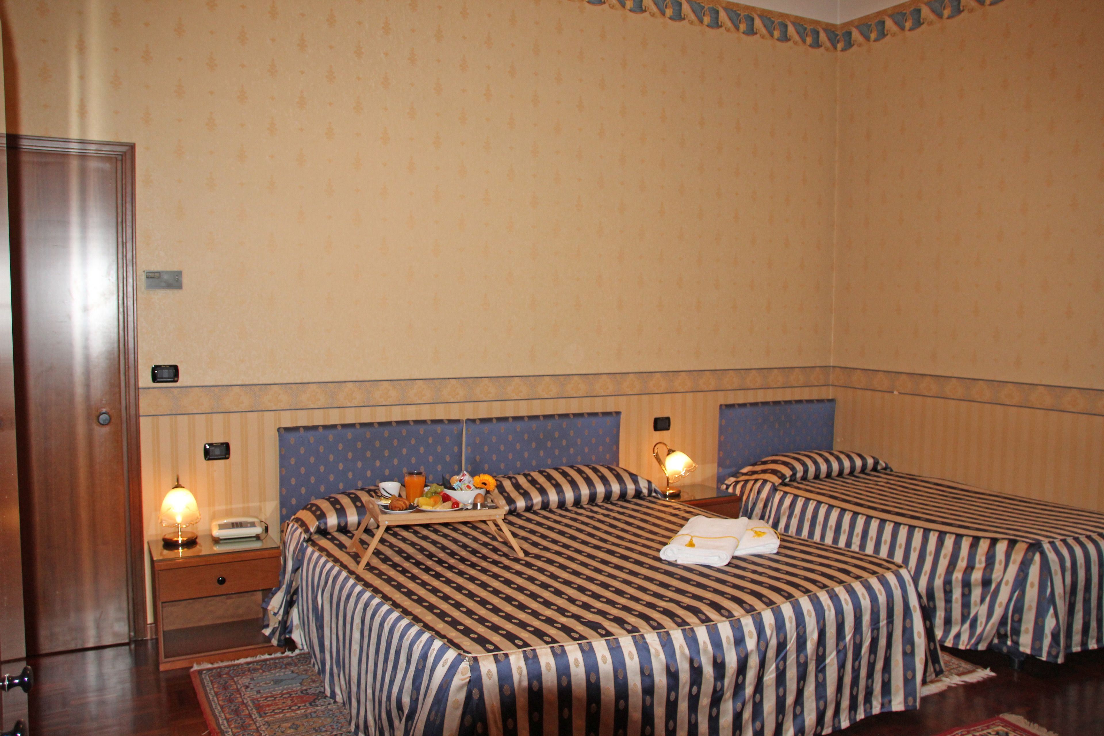 Hotellihuone, jossa on kaksi sänkyä, puinen vaatekaappi, yöpöydät lampuineen ja kuviollinen seinä.