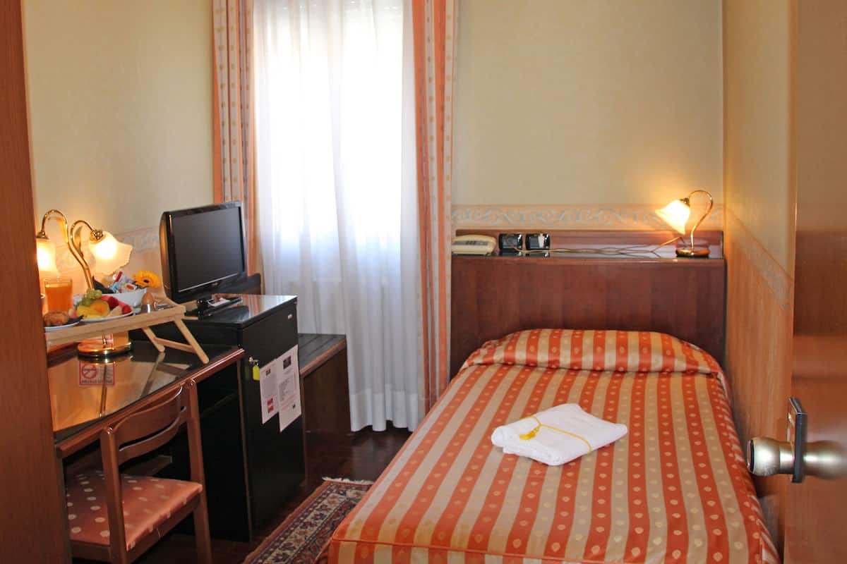A hotel room with a bed, a desk with a computer, a chair, a small table with a lamp, and a window with curtains.