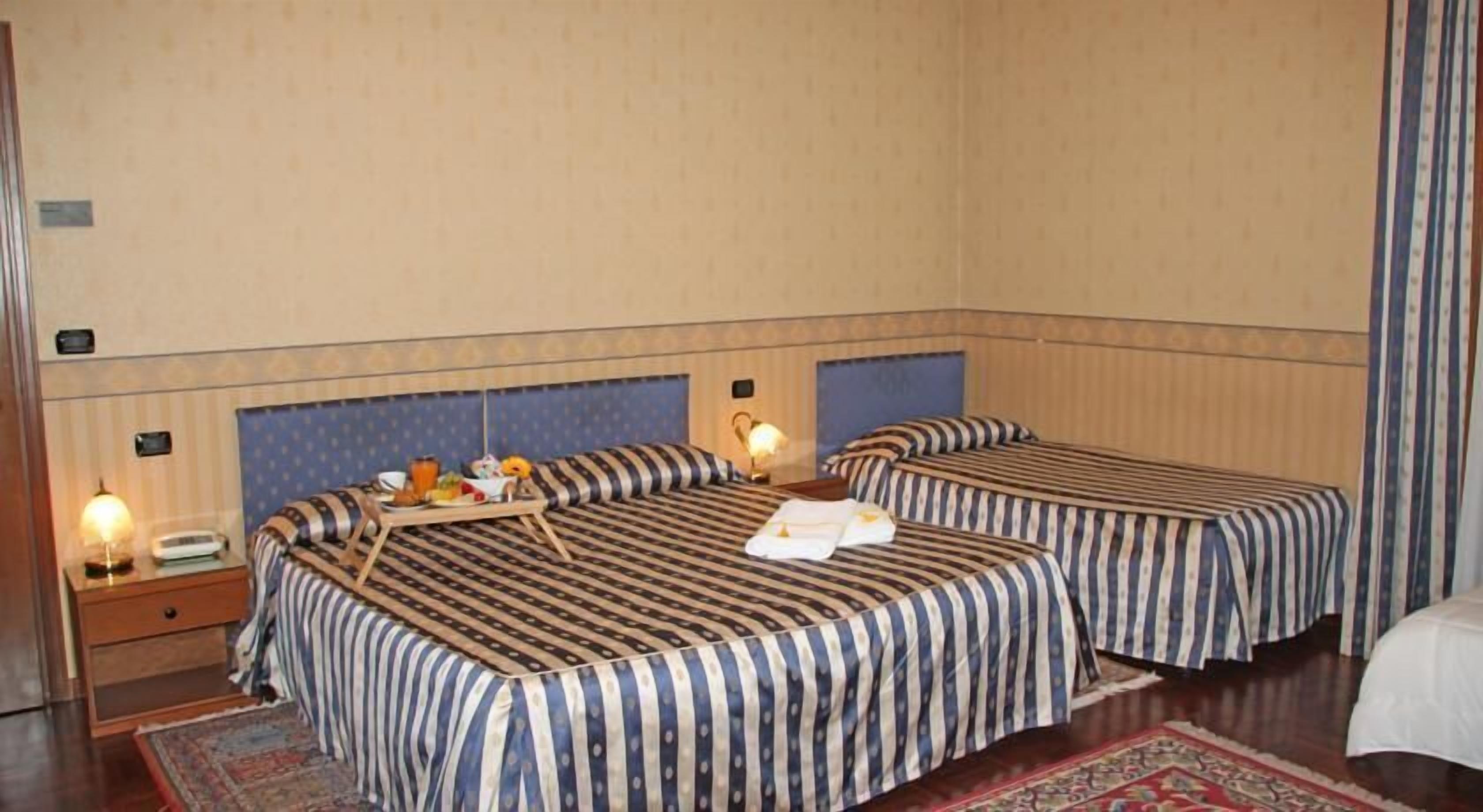 A hotel room with two beds, a bedside table with a lamp, a tray with breakfast items, and a patterned rug.