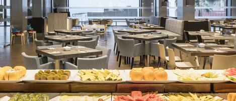 A modern restaurant with multiple dining tables and chairs, a variety of fruits and vegetables displayed on the counter, and large windows offering a view of the city.