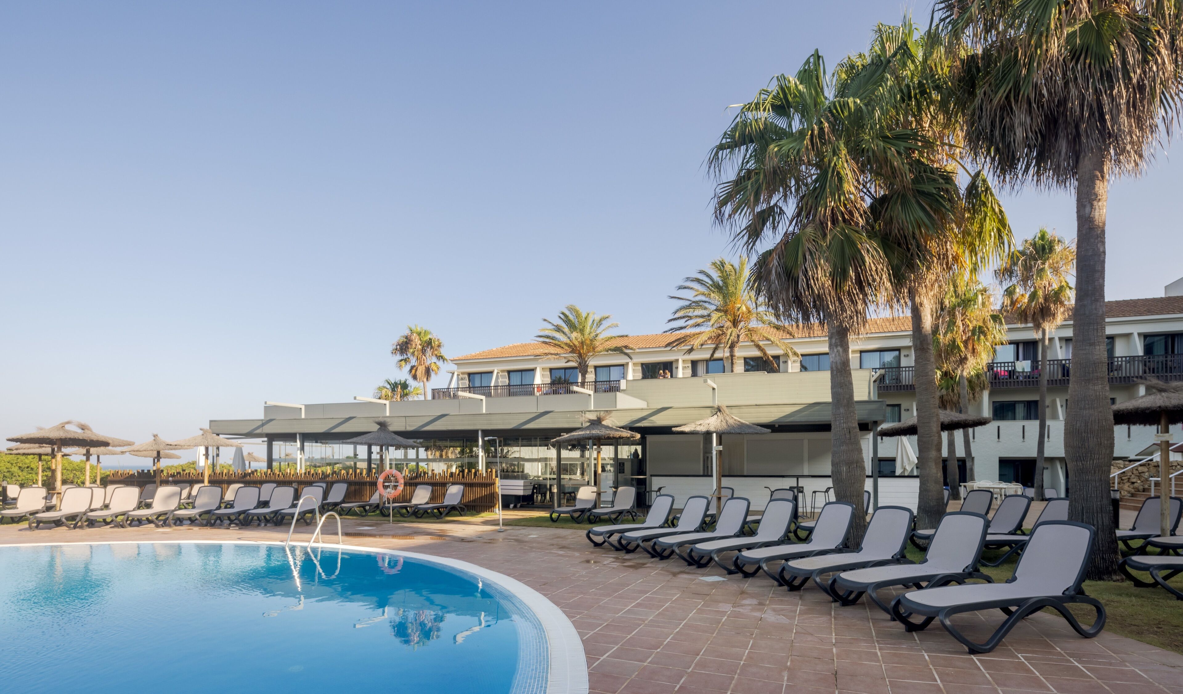 Zona de piscina con sillones de descanso y sombrillas, cielo despejado y un edificio con balcones.