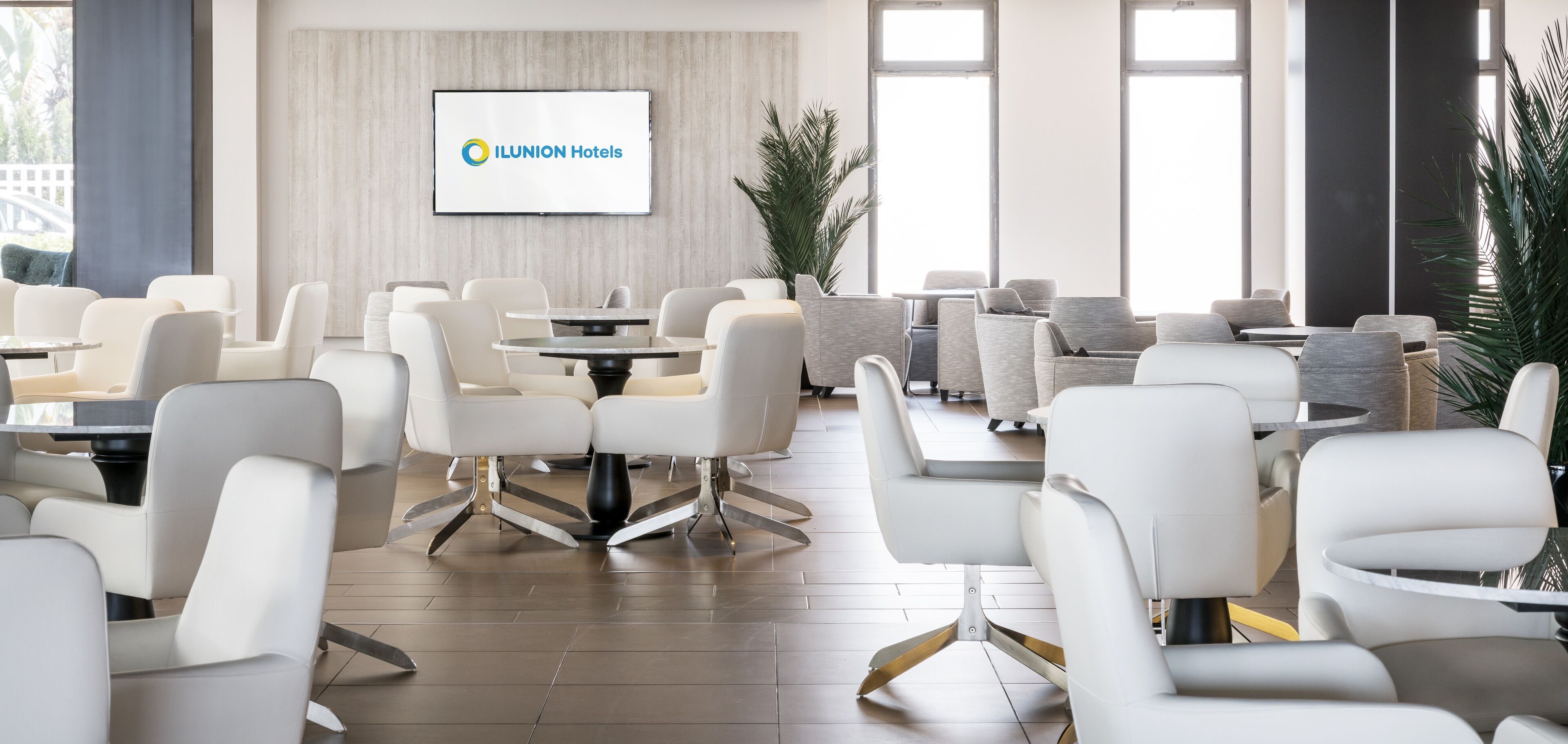 A modern hotel lobby with white furniture, a large screen displaying the hotel name, and large windows.