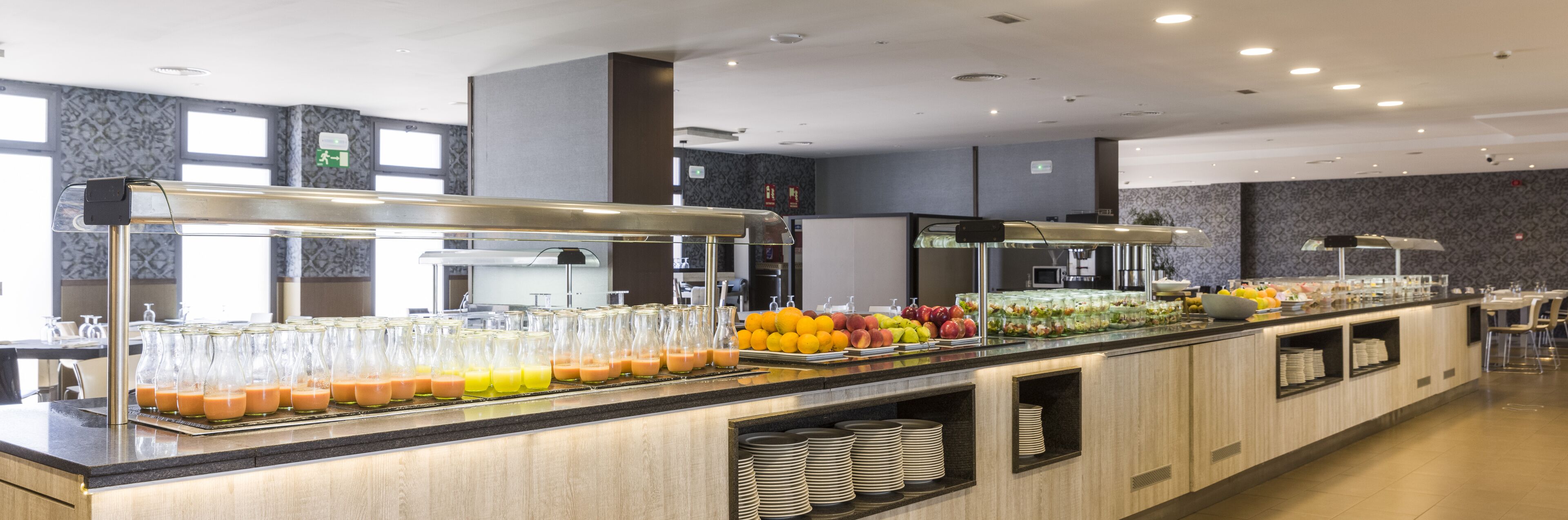 A buffet area with various food items and beverages, including fruits and juices, in a modern setting with a counter and seating area.